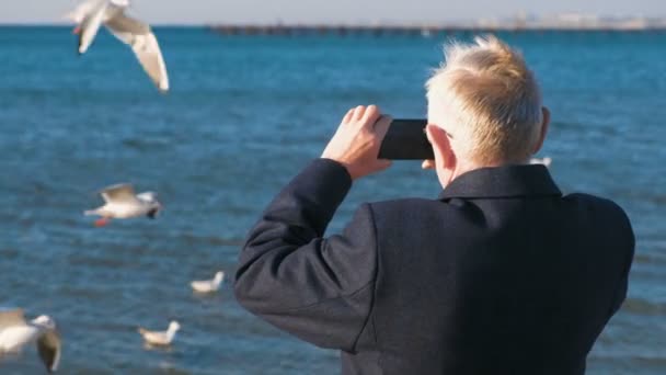 Homme âgé aux cheveux gris photographié sur un paysage marin de téléphone portable avec une jetée et des mouettes. Vue arrière . — Video