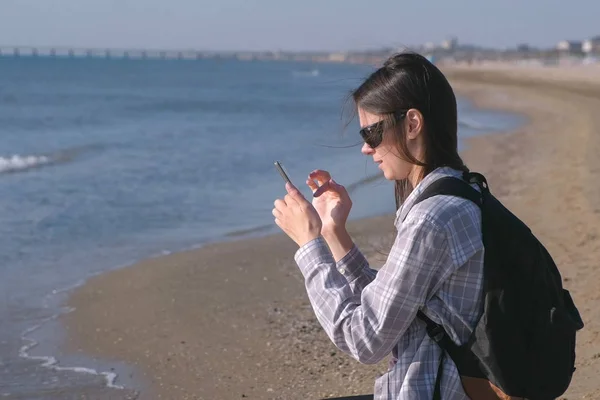 Путешественница девушка с рюкзаком блоггер на песчаном пляже моря и делает фото и видео на мобильный телефон . — стоковое фото