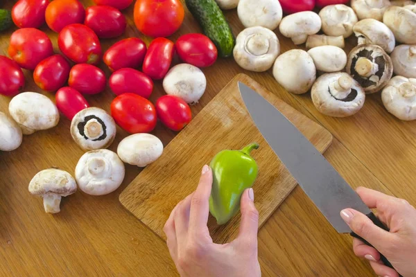 Mujeres manos pelan pimiento verde sobre tabla de madera en la mesa de la cocina con verduras . — Foto de Stock