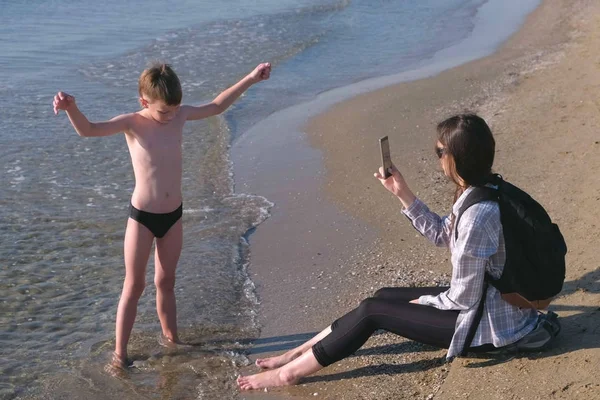 お母さんは彼女の息子の携帯電話で写真やビデオを取ります海沿い歩いて小さなブロガー. — ストック写真