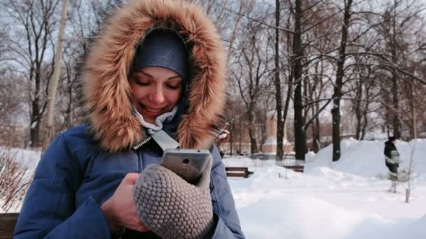 Closeup gelukkige vrouw is zittend op de Bank en browsen van mobiele telefoon in winter park in de stad tijdens de dag in de besneeuwde weer met dalende sneeuw — Stockvideo