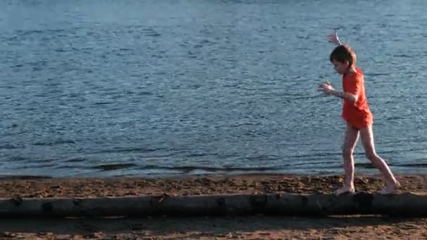Jongen in een rode t-shirt gaat op een log op het strand op de rivier Bank. — Stockvideo