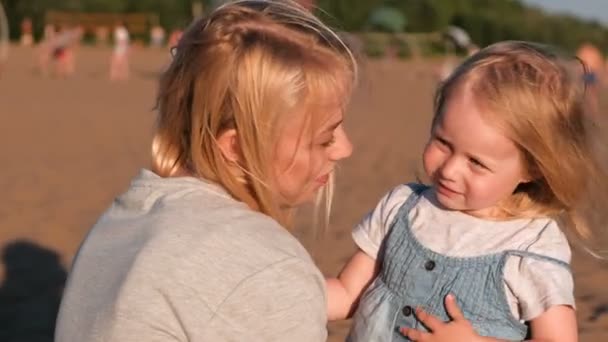 Beautiful blonde mom and daughter cuddling on the beach at sunset. — Stock Video