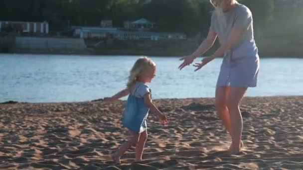 Mama i dziewczyna, gry na plaży nad rzeką o zachodzie słońca. — Wideo stockowe