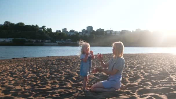 Linda mãe e filha loira feliz abraçando e falando na praia ao pôr do sol . — Vídeo de Stock