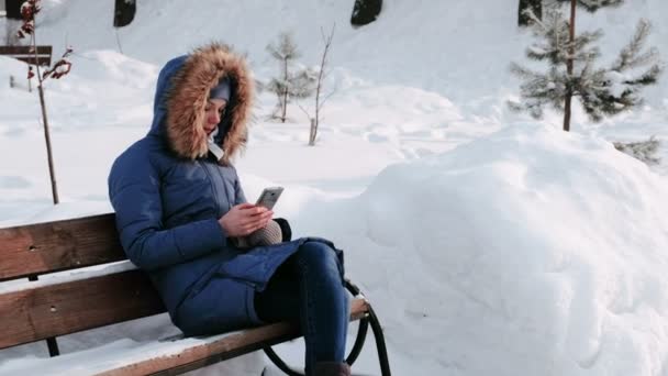 Kvinnan sitter på bänken och surfar mobiltelefon i Vinterparken i staden under dagen i snöiga väder med fallande snö. — Stockvideo