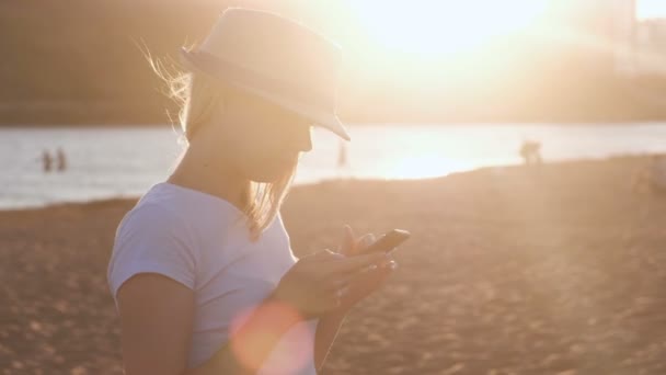 모자에 아름 다운 금발 소녀 해질녘 해변에서 그녀의 휴대 전화에 메시지 유형. — 비디오
