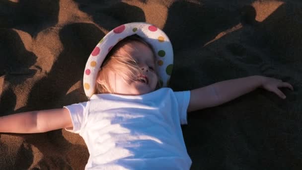 Счастливая девочка блондинка в шляпе, лежащая на пляже на песке . — стоковое видео