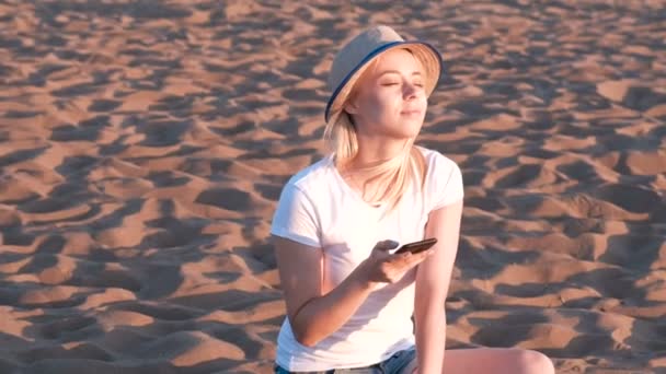 Hermosa chica rubia en un sombrero sentado en la playa al atardecer con teléfono móvil . — Vídeos de Stock