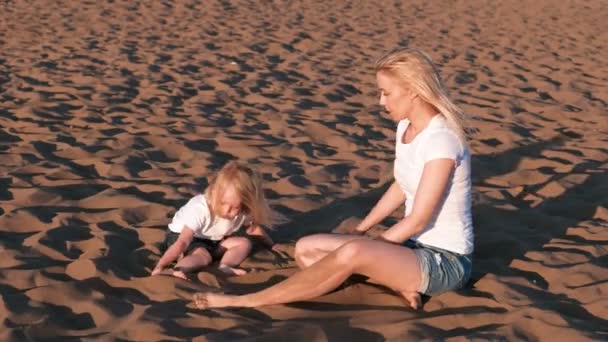 Beautiful blonde mom and daughter play with sand sitting together on the beach. — Stock Video