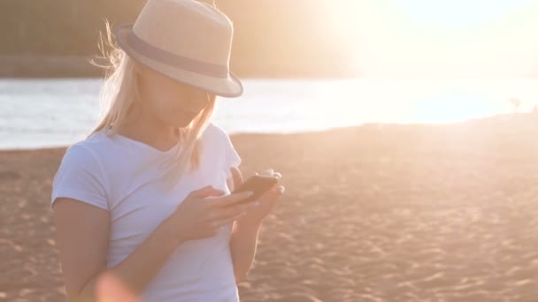 Beautiful blonde girl in a hat types a message on her mobile phone on the beach at sunset. — Stock Video