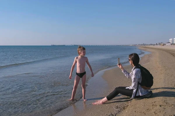 Mamá toma una foto y video en el teléfono móvil de su hijo pequeño blogger caminando por el mar . —  Fotos de Stock