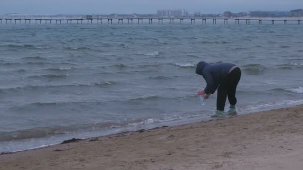 Літня жінка мандрівник отримує пляшку морської води, що стоїть на піщаному пляжі. Осінь на березі моря. Морський сувенір . — стокове відео