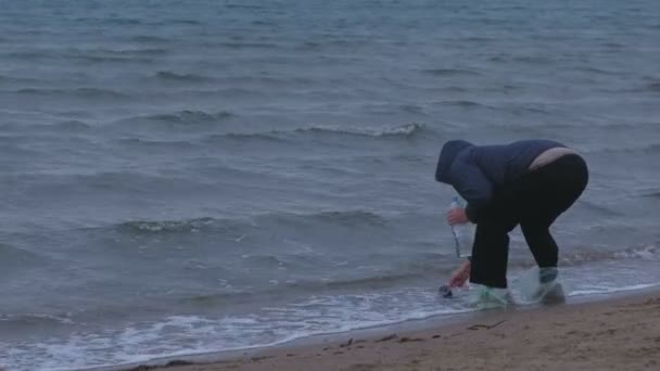 Elderly woman traveler gaining a bottle of sea water standing on a sandy beach. Autumn on the seashore. Sea souvenir. — Stock Video