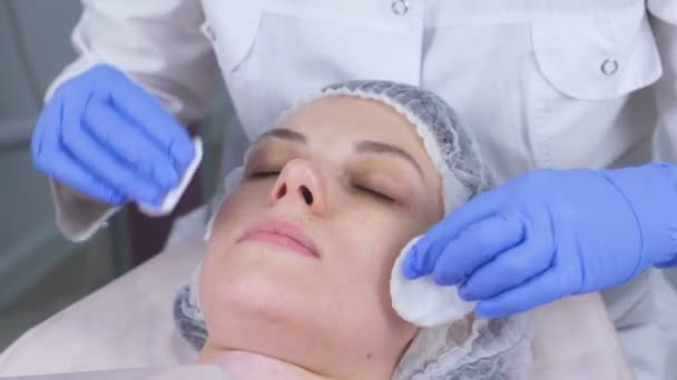 Cosmetologist σκουπίζει το πρόσωπο μιας νεαρής γυναίκας με τα μαξιλάρια βαμβακιού. Τα χέρια του το beautician γάντια και ένα closeup του προσώπου. Θεραπείες ομορφιάς για το πρόσωπο. — Αρχείο Βίντεο