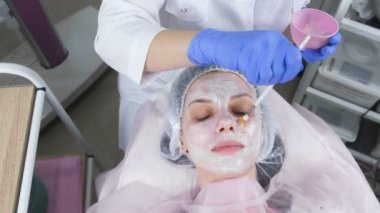 Şevval özçelik güzellik bir fırça ile kadının yüzünde beyaz bir maske koyar. Güzellik uzmanı mavi kauçuk eldiven eller. Yüz bakımları. Üstten görünüm.