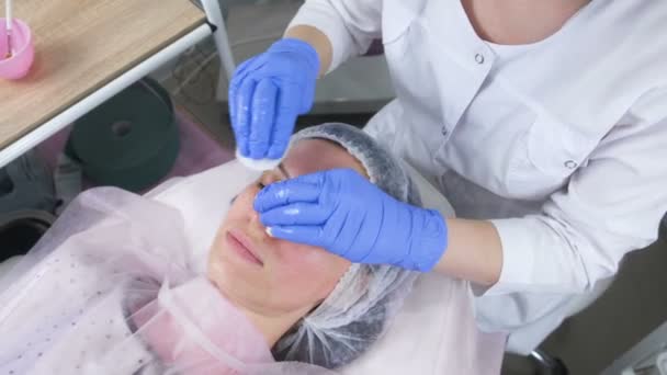 Kosmetolog våtservetter i ansiktet av en ung kvinna med bomullsrondeller. Händerna på kosmetolog i handskar och klienter inför närbild. Ansiktsbehandlingar. — Stockvideo