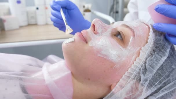 Schoonheidsspecialiste zet een wit masker op het gezicht van de dames met een borstel. De handen van een schoonheidsspecialist in blauwe rubberen handschoenen. Gezichtsbehandelingen. Zijaanzicht. — Stockvideo