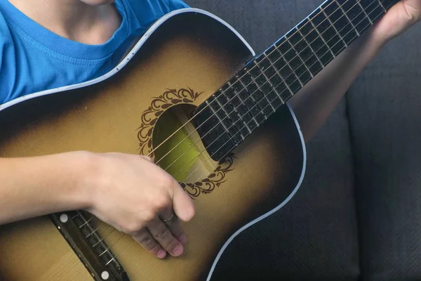 O rapaz está a tocar violão sentado no sofá, com as mãos de perto. Conceito de aprender a tocar um instrumento musical . — Fotografia de Stock