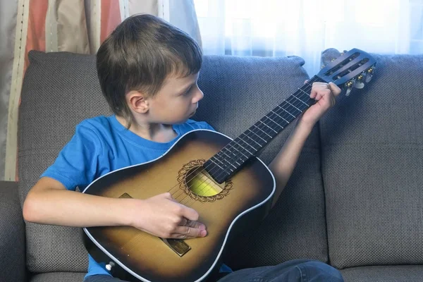 Мальчик учится играть на гитаре, сидя на диване. Концепция обучения игре на музыкальном инструменте . — стоковое фото