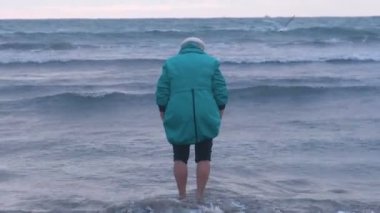 Şapkasını ve ceket yalınayak yaşlı kadın deniz suyu batımında kışın yürüyor. Arkadan görünüm.