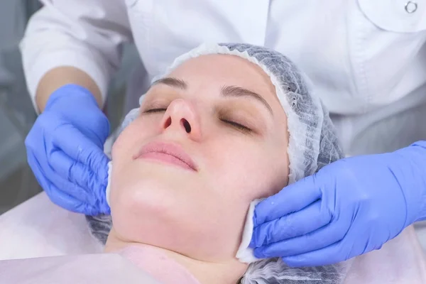 Cosmetologist limpa o rosto de uma jovem mulher com almofadas de algodão. As mãos do esteticista em luvas e uma cara fecham-se. Tratamentos de beleza para o rosto . — Fotografia de Stock