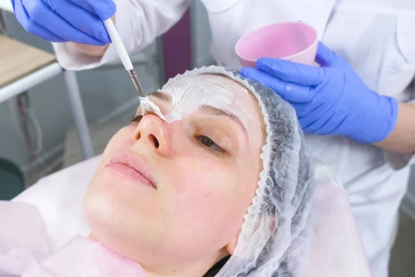 Estetista mette una maschera bianca sul viso delle donne con un pennello. Mani di un cosmetologo in guanti di gomma blu. Trattamenti viso . — Foto Stock