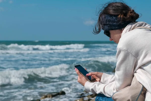Женщина печатает сообщение по телефону, сидя на берегу моря осенью . — стоковое фото