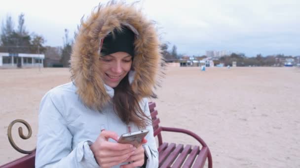 Vrouw in een witte donsjack zittend op een bankje op het strand en het typen van een bericht op sociale media op mobiele telefoon. — Stockvideo