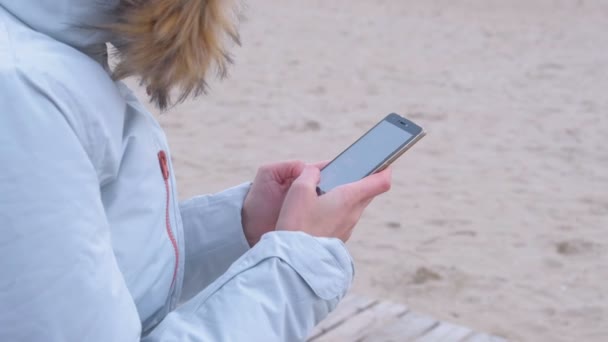Close-up womans manos escribiendo un post en las redes sociales en el teléfono en el fondo del mar. Mar invierno, mujer en chaqueta blanca . — Vídeo de stock