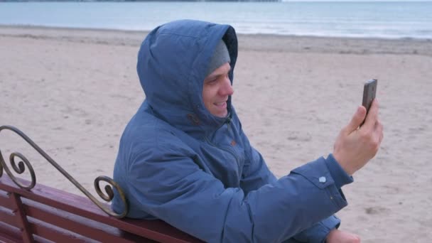 Homem blogueiro em uma jaqueta azul para baixo sentado em um banco na praia de areia e falando um bate-papo por vídeo no telefone celular . — Vídeo de Stock