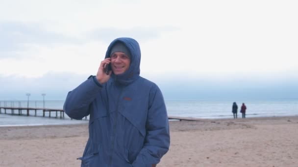 冬天, 穿着夹克在海边的海滩上用手机说话的人快乐的微笑. — 图库视频影像