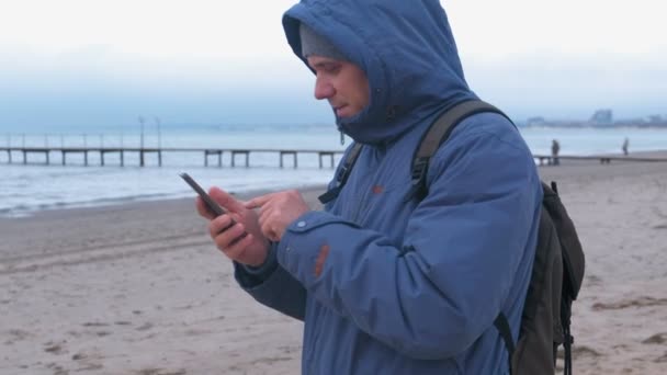 Man in een blauw donsjack aan het zandstrand aan de zee te typen van een bericht op mobiele telefoon. Zijaanzicht. — Stockvideo