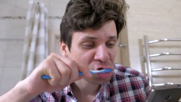 Молодой человек чистит зубы и смотрит на телефон в ванной комнате . — стоковое видео