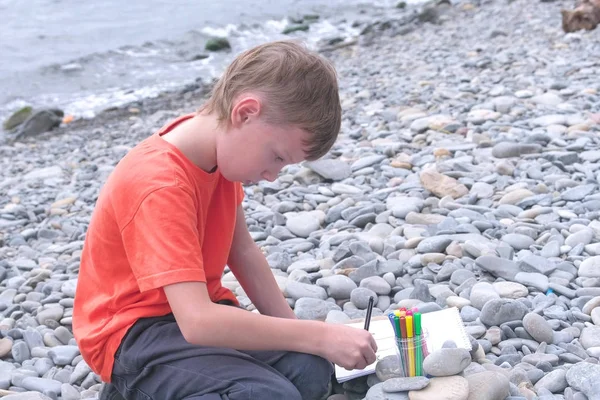 Мальчик рисует маркерами, сидя на каменном пляже на берегу моря — стоковое фото