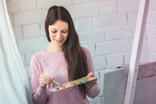 Красивая молодая женщина рисует картину на мольберте. Держит кисть в руках и палитру красок — стоковое фото