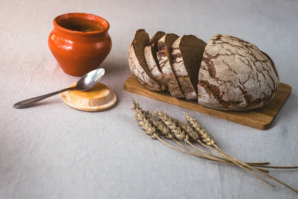 Зерно вкусный домашний хлеб. Кусочки хлеба на разделочной доске. Зерновые уши на льняной скатерти. Горшок с медом и ложкой . — стоковое фото
