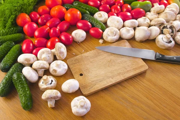 Tablero de madera con cuchillo y verduras frescas tomates, pepinos, calabacines, pimientos, verduras, rábanos y champiñones en la mesa de la cocina . — Foto de Stock