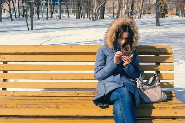 Mulher está navegando na internet em seu telefone sentado no banco no parque da cidade de inverno em dia ensolarado . — Fotografia de Stock