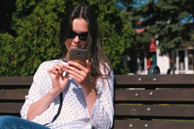 Genç kadın esmer parkta bankta oturmuş cep telefonunda bir mesaj türleri.