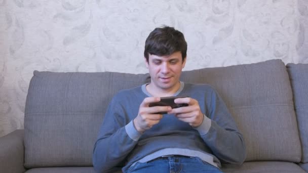 Homem está jogando jogo em seu telefone móvel sentado no sofá . — Vídeo de Stock