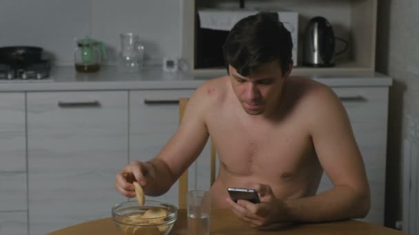 Jonge man het eten van cookies in de keuken bij nacht en telefoon te kijken. Slapeloosheid en angst. Buitensporige voedsel. — Stockvideo