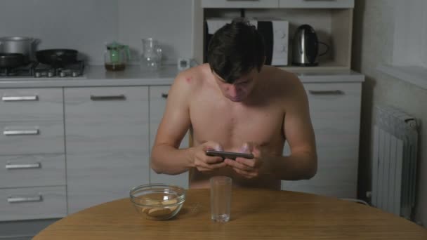 Młody człowiek jedzenie ciasteczek w kuchni w nocy i grania w gry na telefon. Bezsenność i niepokój. Nadmierne jedzenie. — Wideo stockowe