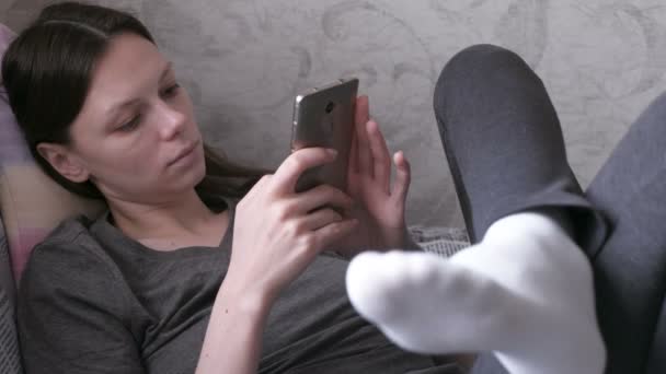 Mujer somnolienta acostada en el sofá y leyendo algo en el teléfono móvil . — Vídeos de Stock