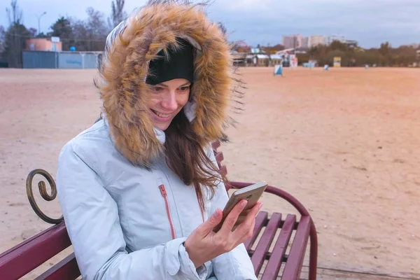 Женщина в белой куртке сидит на скамейке на пляже и смотрит удивительное видео на мобильный телефон . — стоковое фото