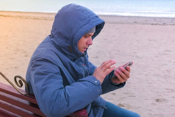 Hombre blogger en una chaqueta azul sentado en un banco en la playa de arena y escribir un post en las redes sociales en el teléfono móvil . — Foto de Stock