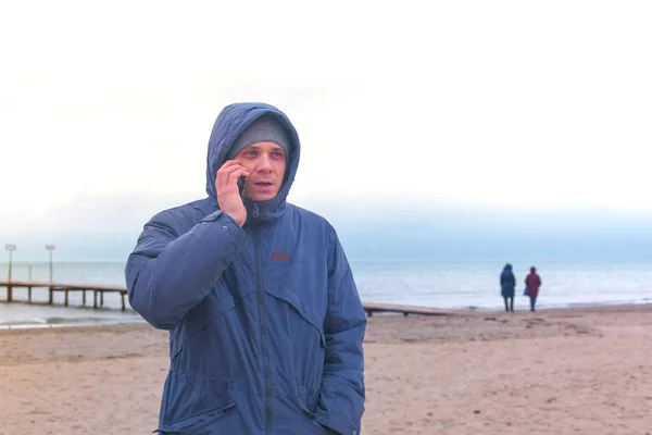 Man i en jacka på stranden vid havet i vinter talar i en mobiltelefon. — Stockfoto