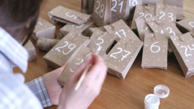 Kadın, çocuklar için Noel varış takvim yapar. Birkaç çanta üzerinde fırça ve beyaz boya ile yazar. Yakın çekim yakışıklı.