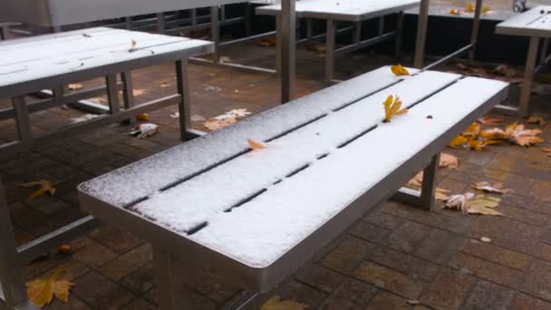 Schneebedeckte Tische und Bänke im Park im Herbst mit gelben Blättern. — Stockvideo