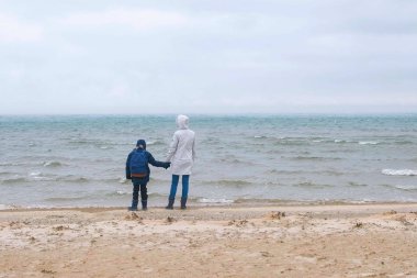 Anne ve oğlu kış, arka görünümünde deniz kum plajda yürüyor. Kum ve kar fırtına güzel manzarasını plaj.